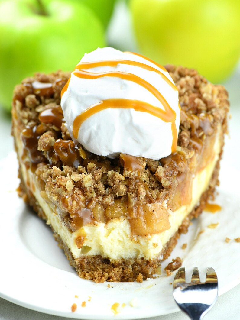 Slice of apple crisp cheesecake pie with a bite taken from it. It features a crumbly oat topping over baked apples and creamy cheesecake filling. The dessert is topped with a dollop of whipped cream and a caramel drizzle, all served on a white plate.