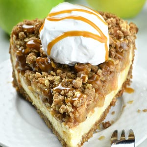 Tempting slice of apple crisp cheesecake pie. It features a crumbly oat topping over baked apples and creamy cheesecake filling. The dessert is topped with a dollop of whipped cream and a caramel drizzle, all served on a white plate.