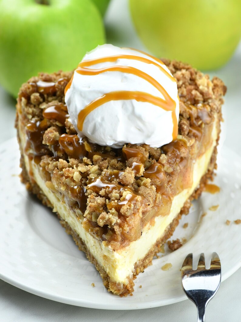 Tempting slice of apple crisp cheesecake pie. It features a crumbly oat topping over baked apples and creamy cheesecake filling. The dessert is topped with a dollop of whipped cream and a caramel drizzle, all served on a white plate.