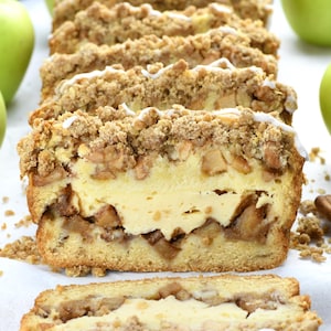 May include: Sliced apple crumble loaf cake with a creamy filling and a streusel topping. The cake is drizzled with white icing. Green apples are in the background. A delicious dessert.
