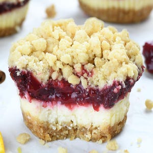 A cranberry mini cheesecake with visible layers of graham cracker crust, creamy cheesecake, tangy cranberry filling, and a golden streusel topping, set on parchment paper with cinnamon sticks and citrus nearby.