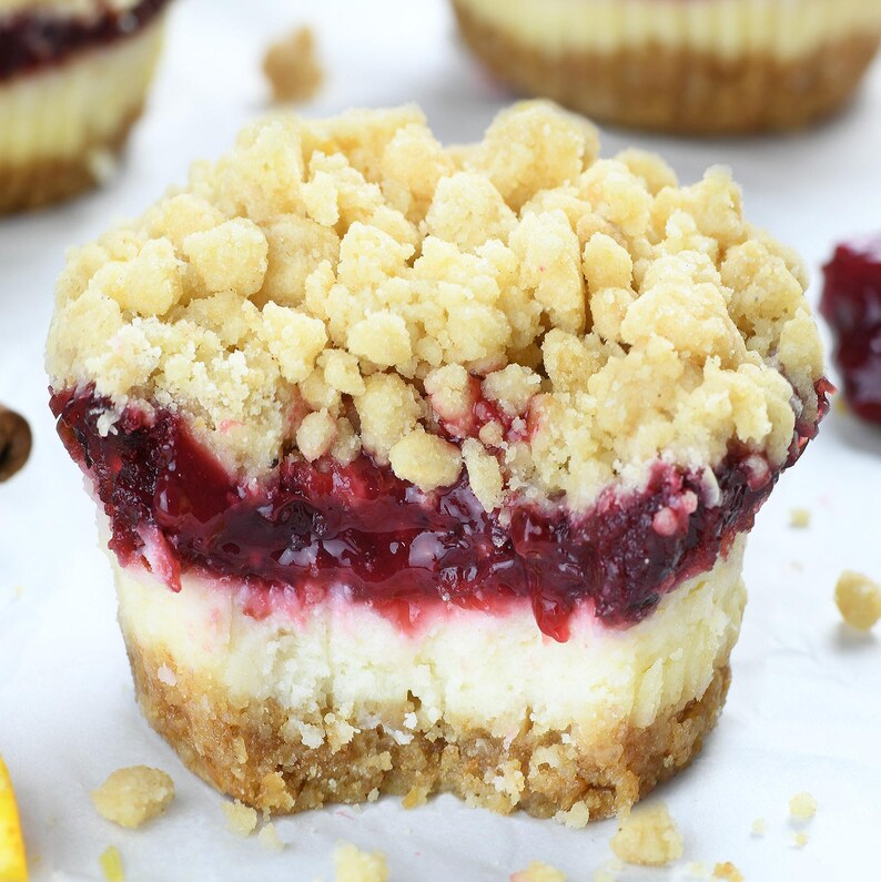 A cranberry mini cheesecake with visible layers of graham cracker crust, creamy cheesecake, tangy cranberry filling, and a golden streusel topping, set on parchment paper with cinnamon sticks and citrus nearby.