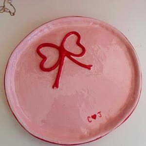 May include: A pink ceramic trinket dish with a red bow design in the centre. The dish has a raised edge and the initials "C ❤️ J" are painted on the side. The bow is a darker shade of red than the edge.
