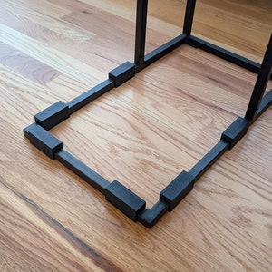 May include: Black metal side table with a square top and four black rubber feet. The table is on a wooden floor.