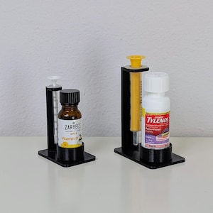 Two black stands, each with a cylinder holding a small medicine bottle and ataller stand behind holding a small syringe