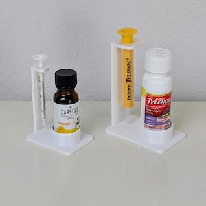 May include: Two white plastic dispensers with clear syringes and bottles of liquid medicine. The bottle on the left is labelled "Zarbee's Baby Vitamin D". The bottle on the right is labelled "Infants' Tylenol Pain + Fever Grape".