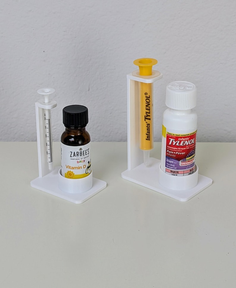 May include: Two white plastic dispensers with clear syringes and bottles of liquid medicine. The bottle on the left is labelled "Zarbee's Baby Vitamin D". The bottle on the right is labelled "Infants' Tylenol Pain + Fever Grape".