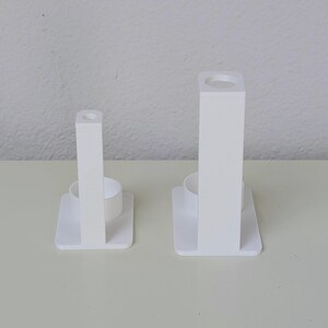 May include: Two white plastic toothbrush holders, each with a small round opening at the top and a larger round opening at the bottom. The holders are of different sizes.