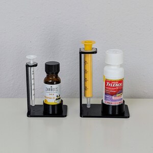 May include: Three black plastic stands with clear glass bottles and a yellow syringe. The bottles are labelled "Zarbee's Baby Vitamin D" and "Infant Tylenol Pain & Fever Grape"