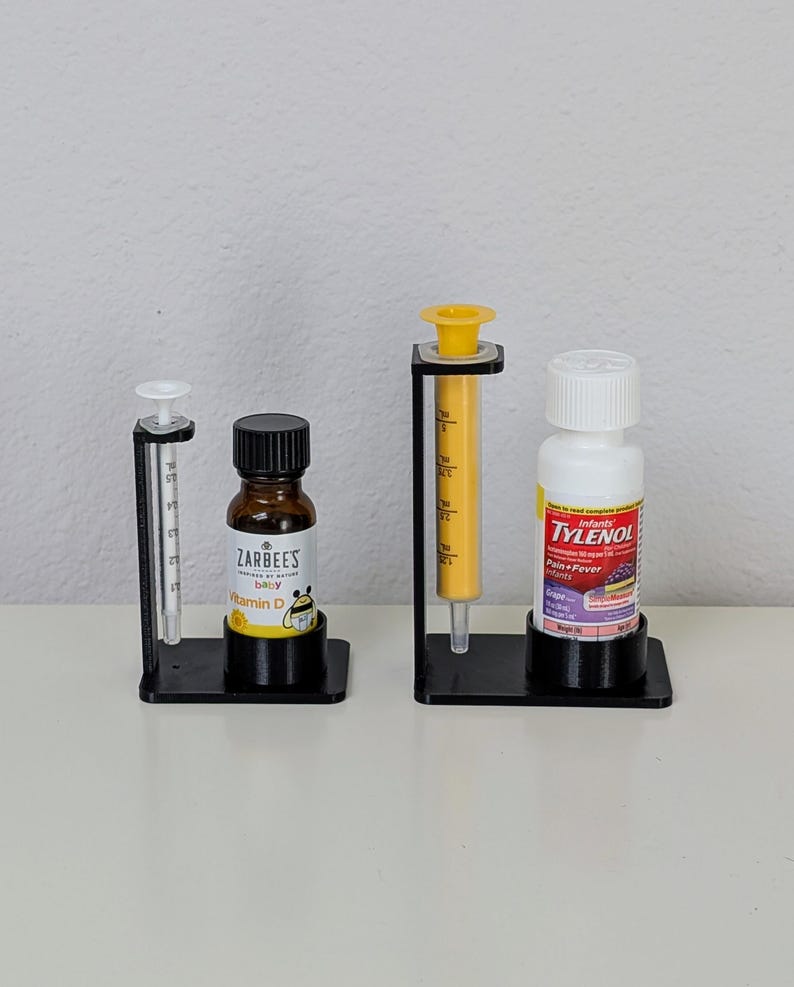 May include: Three black plastic stands with clear glass bottles and a yellow syringe. The bottles are labelled "Zarbee's Baby Vitamin D" and "Infant Tylenol Pain & Fever Grape"