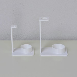May include: Two white plastic toothbrush holders with a small cup for toothpaste. The holders are different sizes.