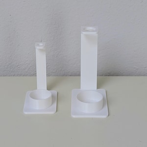 May include: Two white plastic toothbrush holders with a round base and a tall, rectangular stand. The holder on the right is taller than the holder on the left.