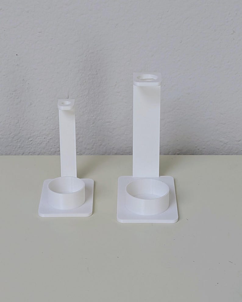 May include: Two white plastic toothbrush holders with a round base and a tall, rectangular stand. The holder on the right is taller than the holder on the left.