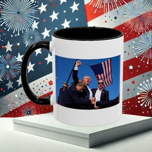 May include: A white coffee mug with a black rim and handle. The mug features a photo of a group of people, including one person raising their fist in the air. The photo is set against a blue sky with a waving American flag in the background.