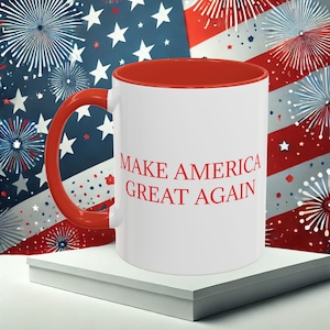 May include: White ceramic mug with a red interior and handle. The mug features the text "MAKE AMERICA GREAT AGAIN" in red. The background includes a stylized American flag and fireworks.