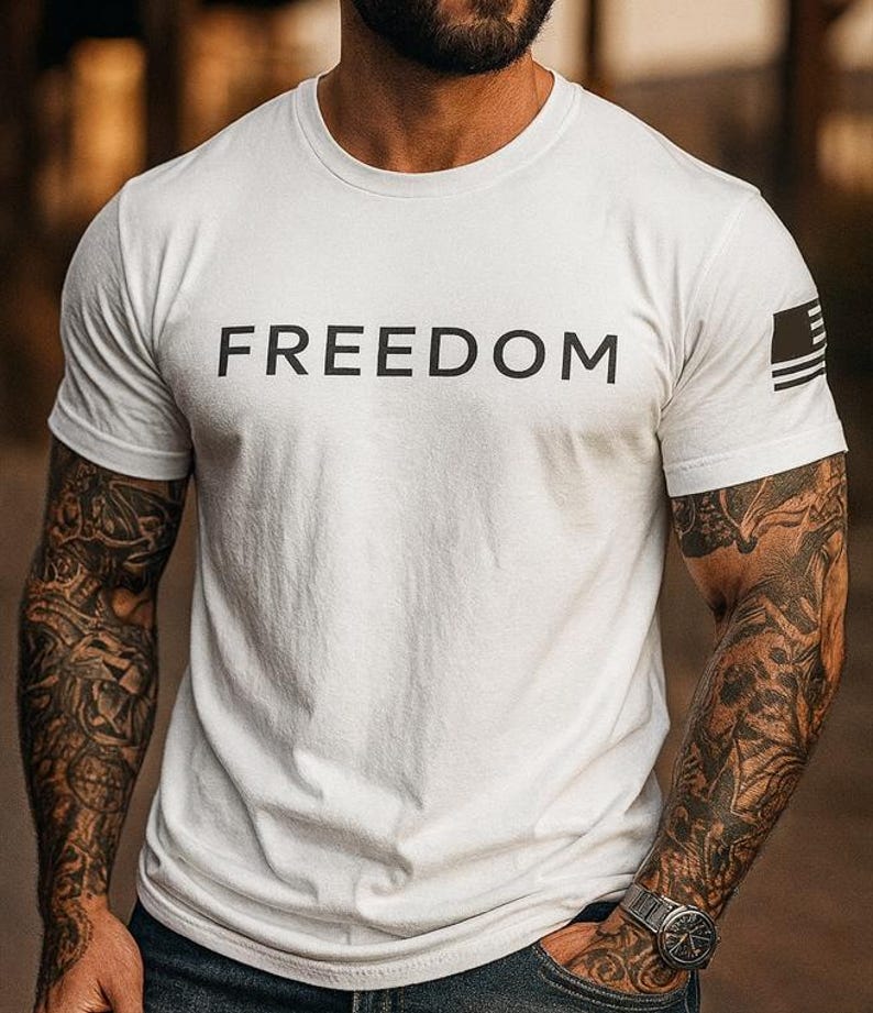 May include: White crew neck t-shirt with the word "FREEDOM" printed in black across the chest. The right sleeve features a small black and white flag design. The shirt is short-sleeved.