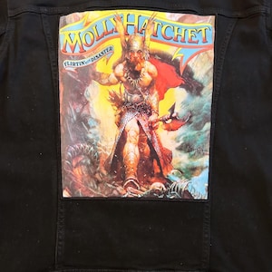 Op de afbeelding: Zwarte spijkerjas met een grote, kleurrijke patch op de rug. De patch toont de bandnaam "Molly Hatchet" en de woorden "Flirtin' with Disaster" boven een illustratie van een krijger met een bijl.
