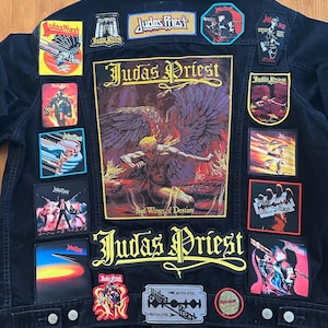 Puede incluir: Chaqueta vaquera negra adornada con múltiples parches de la banda Judas Priest. Los parches muestran arte de álbumes y el logotipo de la banda en varios colores. La chaqueta tiene botones plateados y un diseño clásico de mezclilla.