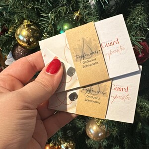 May include: Two small white and tan product cards are held in front of a Christmas tree. The cards have text that reads "tropfenweise OnGuard Zahnpasta" and "Guard upasta". The background is a green Christmas tree with gold and red ornaments.