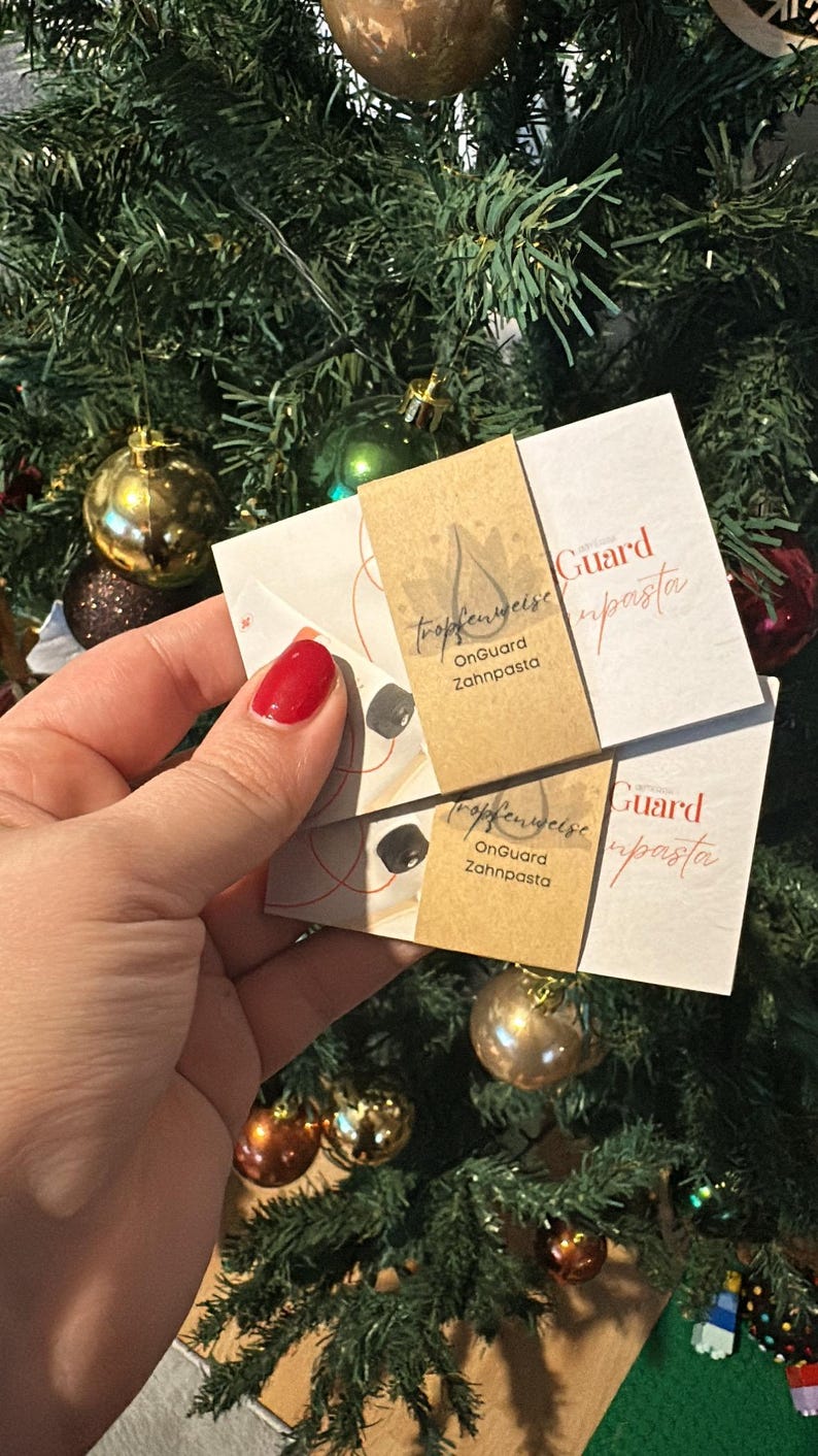 May include: Two small white and tan product cards are held in front of a Christmas tree. The cards have text that reads "tropfenweise OnGuard Zahnpasta" and "Guard upasta". The background is a green Christmas tree with gold and red ornaments.