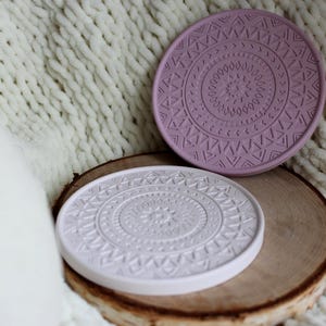 Könnte beinhalten: Zwei runde Untersetzer mit geprägtem geometrischem Design. Einer ist weiß, der andere lila. Sie zeigen ein detailliertes Muster. Sie liegen auf einer Holzscheibe, vor einem weichen, strukturierten Hintergrund.