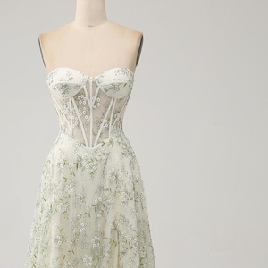 May include: A white strapless gown with a fitted bodice and a flowing skirt. The bodice features a sheer panel with delicate floral embroidery. The skirt is adorned with a floral pattern in shades of green.