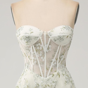 May include: A white strapless dress with a floral pattern and a corset-style bodice. The dress is made of a sheer fabric and has a fitted silhouette.