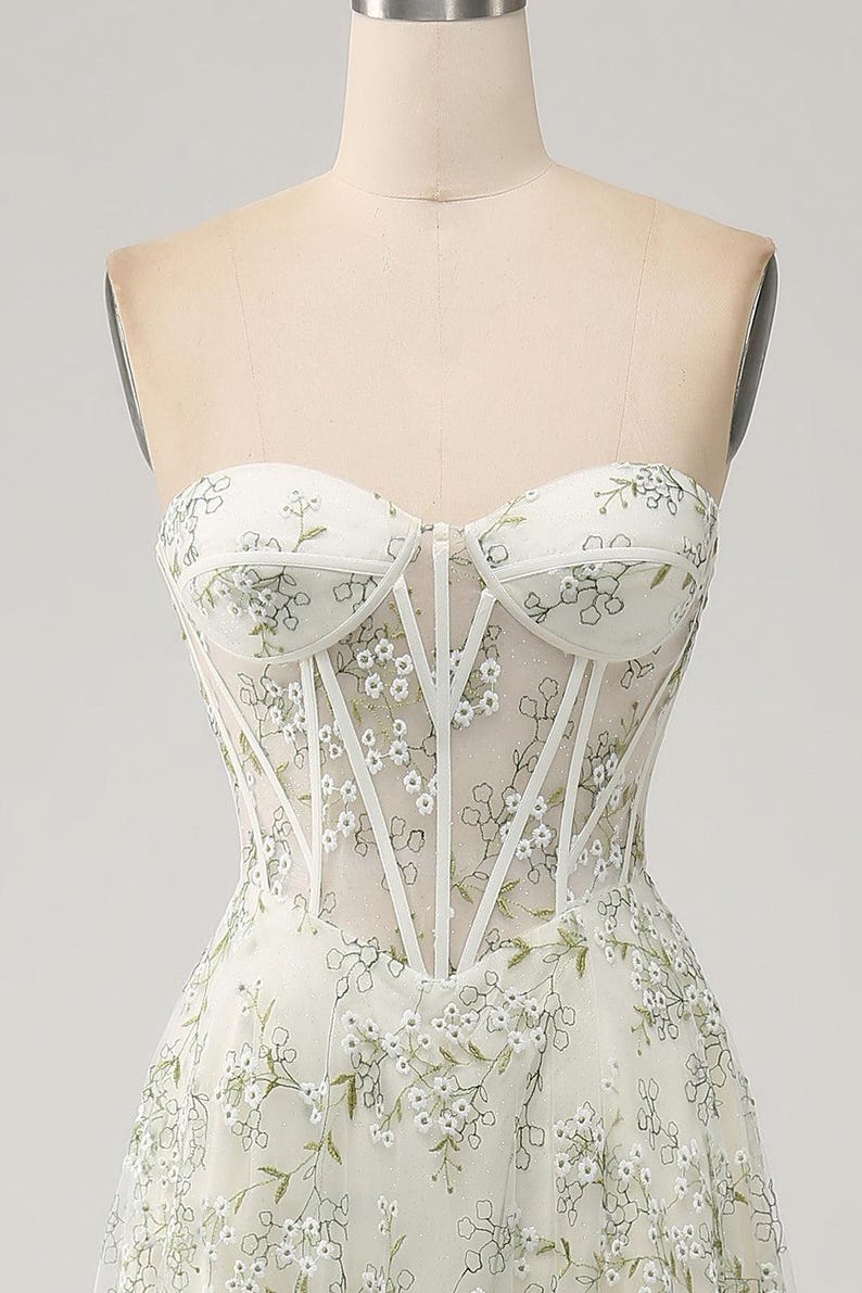 May include: A white strapless dress with a floral pattern and a corset-style bodice. The dress is made of a sheer fabric and has a fitted silhouette.
