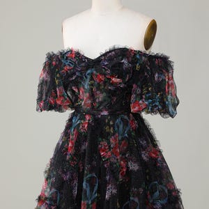 May include: A black off-the-shoulder dress with a floral pattern in red, blue, and purple. The dress has a ruffled neckline and sleeves, and a fitted waist. The skirt is flared and has ruffles.
