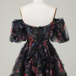May include: Black off-the-shoulder dress with a floral pattern. The dress has a ruffled neckline and sleeves, and a layered skirt. The floral print includes pink, red, blue, and purple flowers. The dress is displayed on a mannequin.