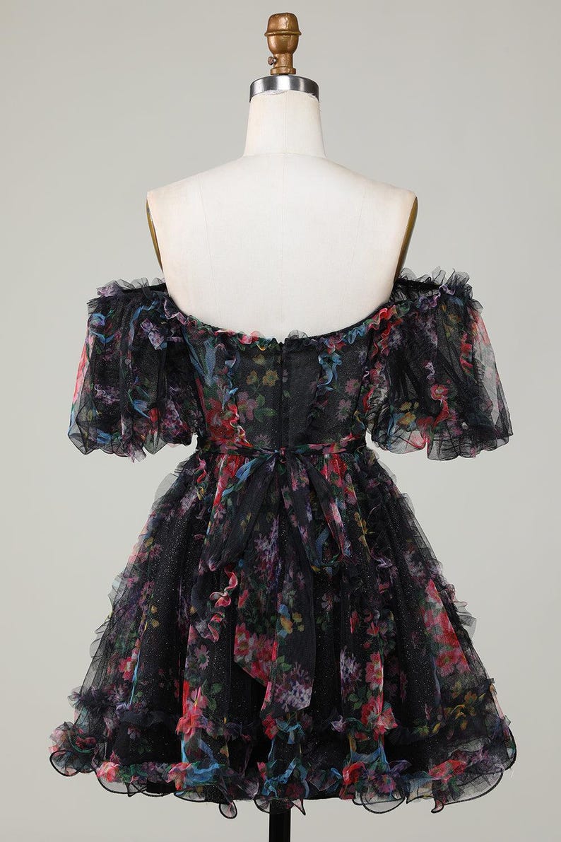 May include: Black off-the-shoulder dress with a floral pattern. The dress has a ruffled neckline and sleeves, and a layered skirt. The floral print includes pink, red, blue, and purple flowers. The dress is displayed on a mannequin.