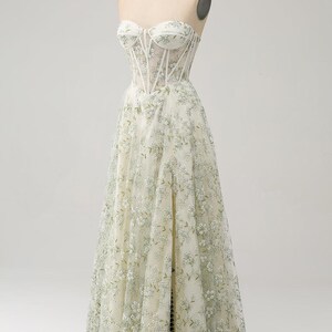 May include: A white strapless gown with a fitted bodice and a flowing skirt. The bodice features a sheer mesh overlay with delicate floral embroidery. The skirt is adorned with a similar floral pattern.