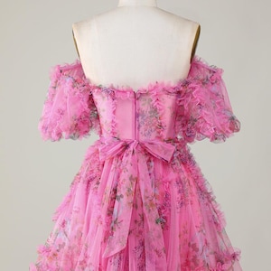 May include: A pink, off-the-shoulder dress with a floral pattern. The dress has ruffled sleeves and a tiered skirt. A bow detail is at the waist. The dress is displayed on a mannequin.