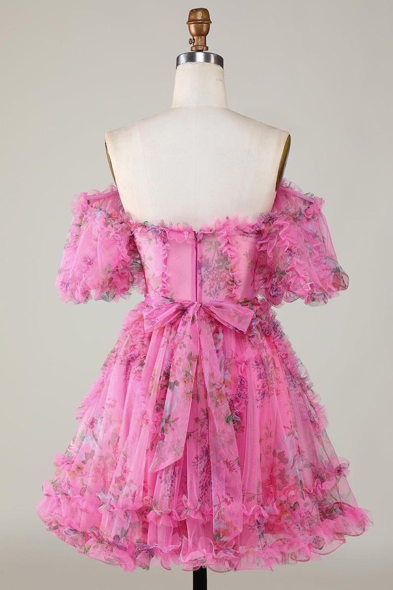 May include: A pink, off-the-shoulder dress with a floral pattern. The dress has ruffled sleeves and a tiered skirt. A bow detail is at the waist. The dress is displayed on a mannequin.