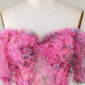 May include: A pink, off-the-shoulder dress with a floral pattern and ruffled trim. The dress is displayed on a mannequin and features a sweetheart neckline and a fitted waist. The fabric appears to be a sheer, lightweight material.