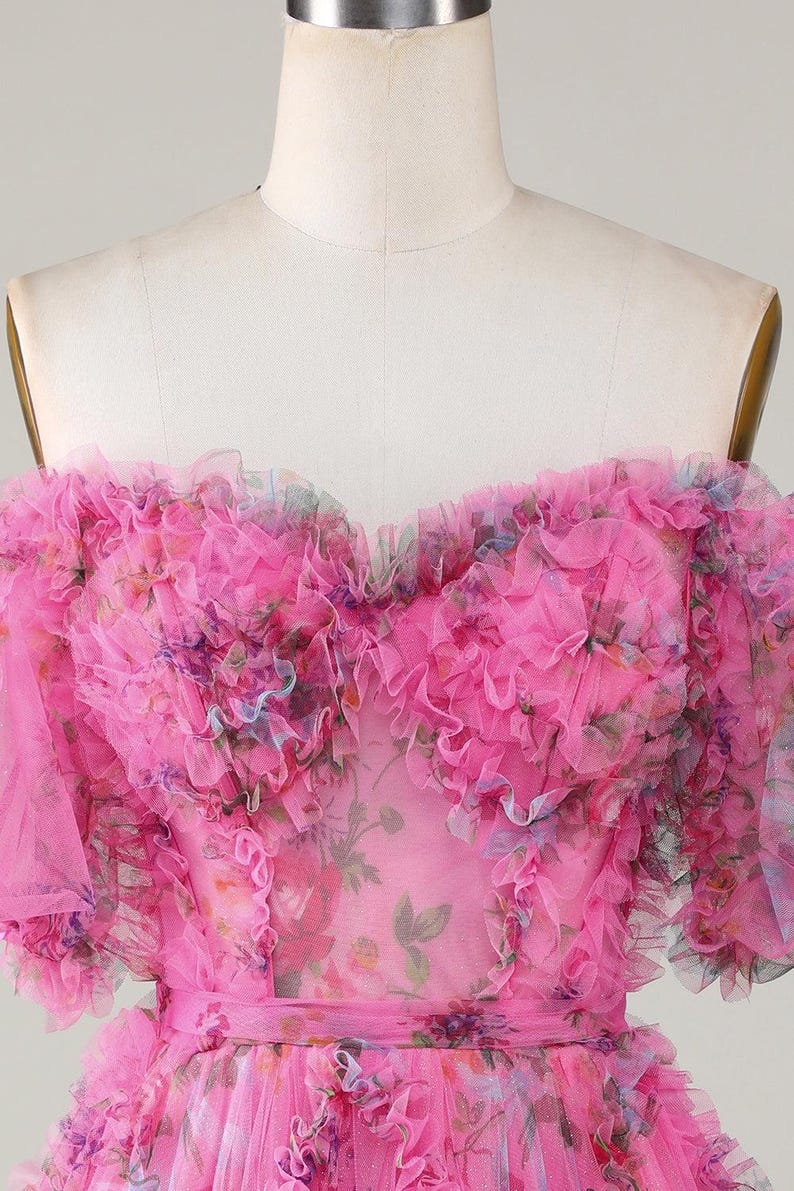 May include: A pink, off-the-shoulder dress with a floral pattern and ruffled trim. The dress is displayed on a mannequin and features a sweetheart neckline and a fitted waist. The fabric appears to be a sheer, lightweight material.