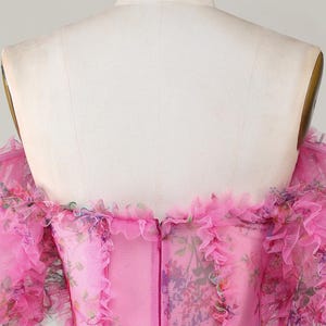 May include: A pink, off-the-shoulder dress with floral print and ruffled trim. The dress features a fitted bodice, a bow at the waist, and a zip closure down the back. The fabric has a sheer, layered appearance.