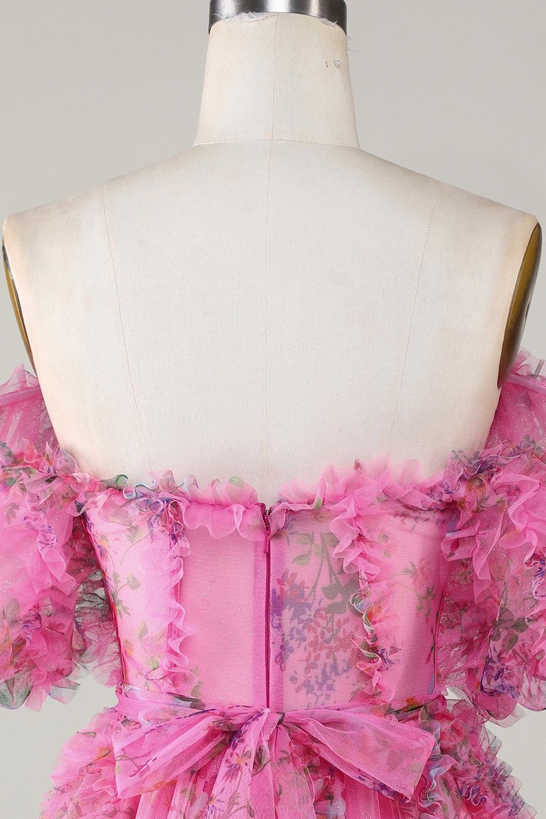 May include: A pink, off-the-shoulder dress with floral print and ruffled trim. The dress features a fitted bodice, a bow at the waist, and a zip closure down the back. The fabric has a sheer, layered appearance.