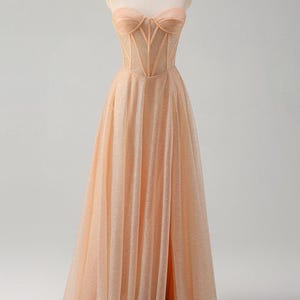 May include: A peach-coloured strapless gown with a fitted bodice and a flowing skirt. The bodice features a sheer mesh panel with delicate stitching. The skirt has a high slit and is adorned with shimmering sequins.