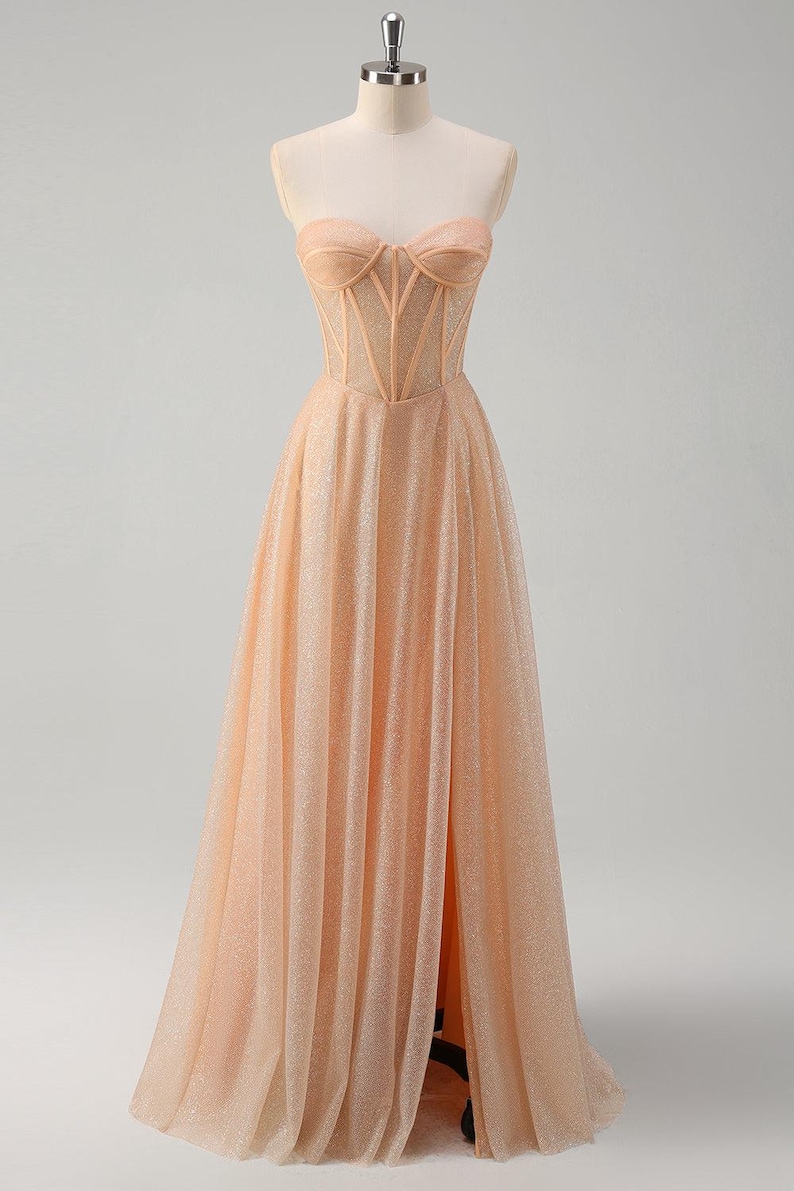 May include: A peach-coloured strapless gown with a fitted bodice and a flowing skirt. The bodice features a sheer mesh panel with delicate stitching. The skirt has a high slit and is adorned with shimmering sequins.