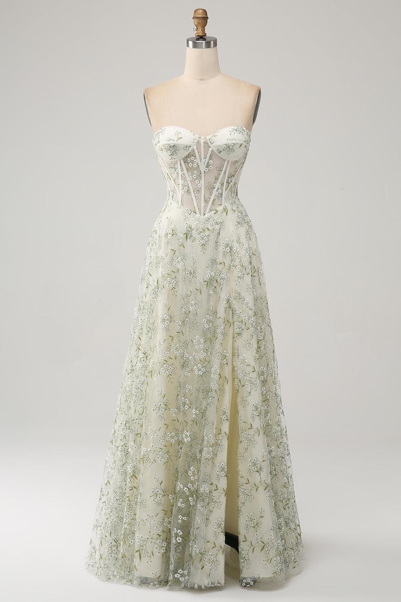 May include: A white strapless gown with a fitted bodice and a flowing skirt. The bodice features a sheer panel with delicate floral embroidery. The skirt is adorned with a floral pattern in shades of green.