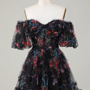 May include: Black off-the-shoulder dress with a floral pattern in red, blue, and purple. The dress has a ruffled neckline, sleeves, and hem. The dress is displayed on a mannequin.