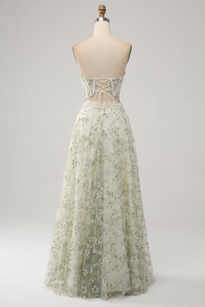 May include: A strapless, floor-length gown with a fitted bodice and a flowing skirt. The bodice is made of a sheer fabric with floral embroidery in shades of green and white. The skirt is made of a soft, flowing tulle fabric.