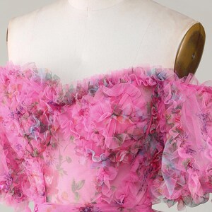 May include: A bright pink, off-the-shoulder dress with layers of ruffled tulle. The dress has a floral print in pink, blue, and purple. The bodice is fitted, and the waist is cinched with a matching belt. A romantic, feminine design.