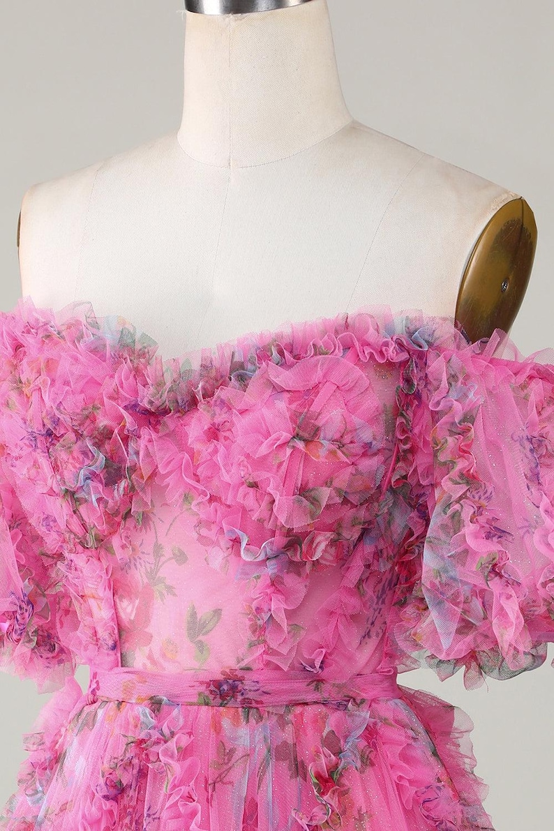 May include: A bright pink, off-the-shoulder dress with layers of ruffled tulle. The dress has a floral print in pink, blue, and purple. The bodice is fitted, and the waist is cinched with a matching belt. A romantic, feminine design.