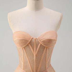 May include: A strapless, champagne-coloured gown with a fitted bodice and a flowing skirt. The bodice features a mesh overlay with a corset-style design.