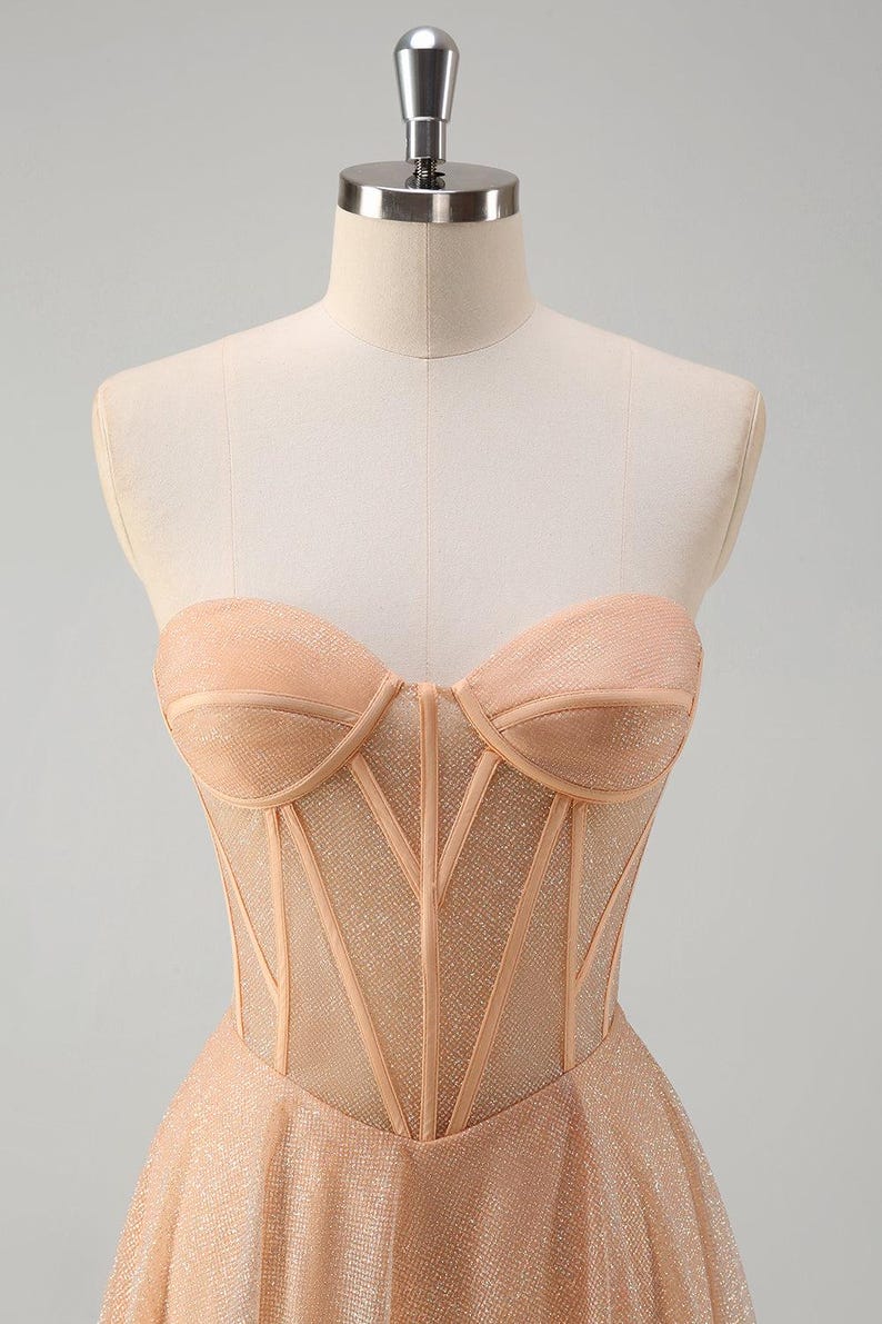 May include: A strapless, champagne-coloured gown with a fitted bodice and a flowing skirt. The bodice features a mesh overlay with a corset-style design.