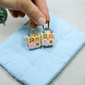 Peut inclure: Une paire de boucles d'oreilles en perles en forme de petits sacs à main avec une poignée rayée noir et blanc et un motif floral coloré.