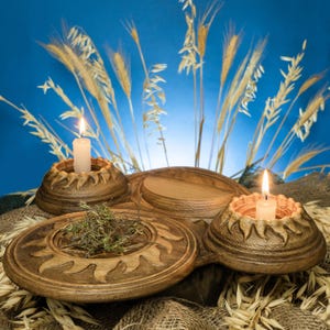 Puede incluir: Un portavelas de madera con tres secciones, dos de ellas con velas encendidas. La sección central contiene hierbas secas. El soporte está decorado con un diseño de sol y colocado sobre un fondo azul con espigas de trigo.