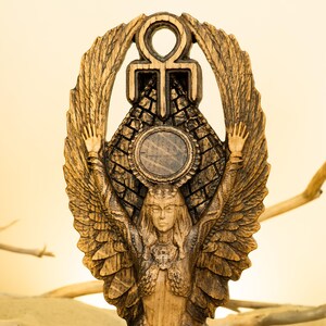 Może przedstawiać: Drewniana rzeźba skrzydlatego stworzenia z rozpostartymi ramionami, trzymającego symbol ankh nad piramidą. Postać otoczona jest okrągłą aureolą i ma szczeg&oacute;łową twarz.